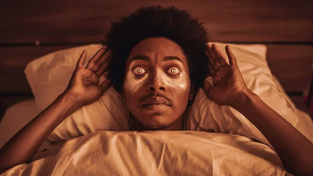 Person with wide eyes in bed, hands near head.